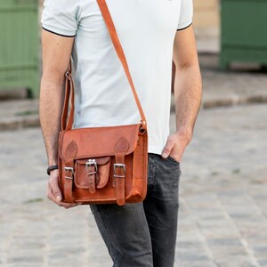 May include: A man wearing a light blue polo shirt with a brown leather satchel bag slung over his shoulder. He is wearing dark blue jeans and white trainers.