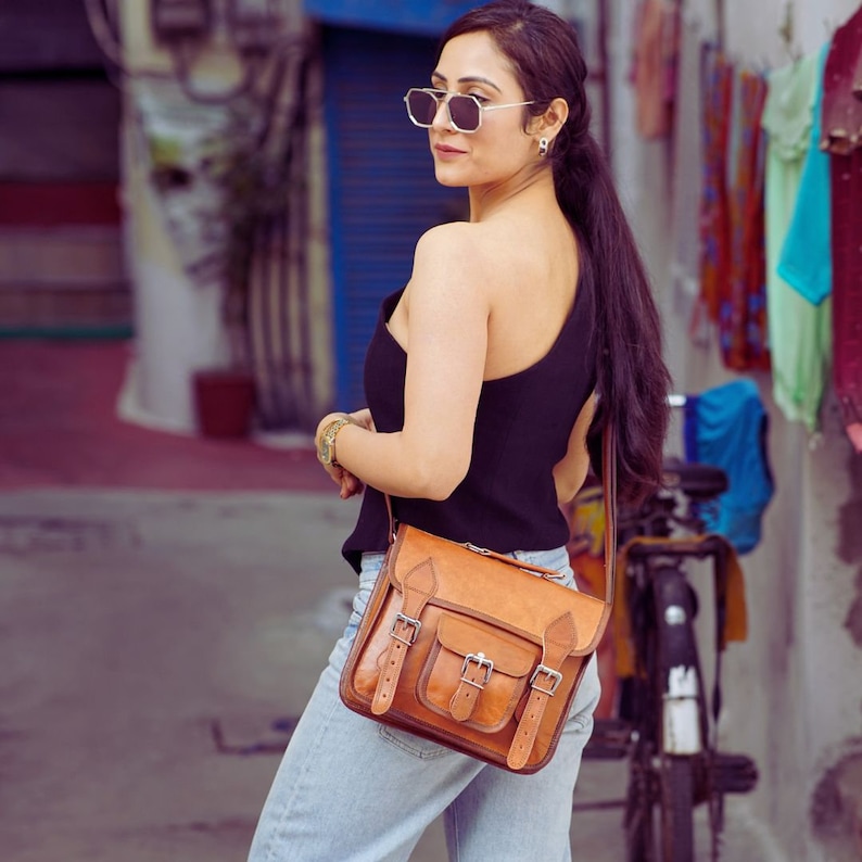 May include: A woman wearing a black one-shoulder top and blue jeans carries a brown leather satchel bag with multiple buckles. The bag has a long strap and is worn over the shoulder.