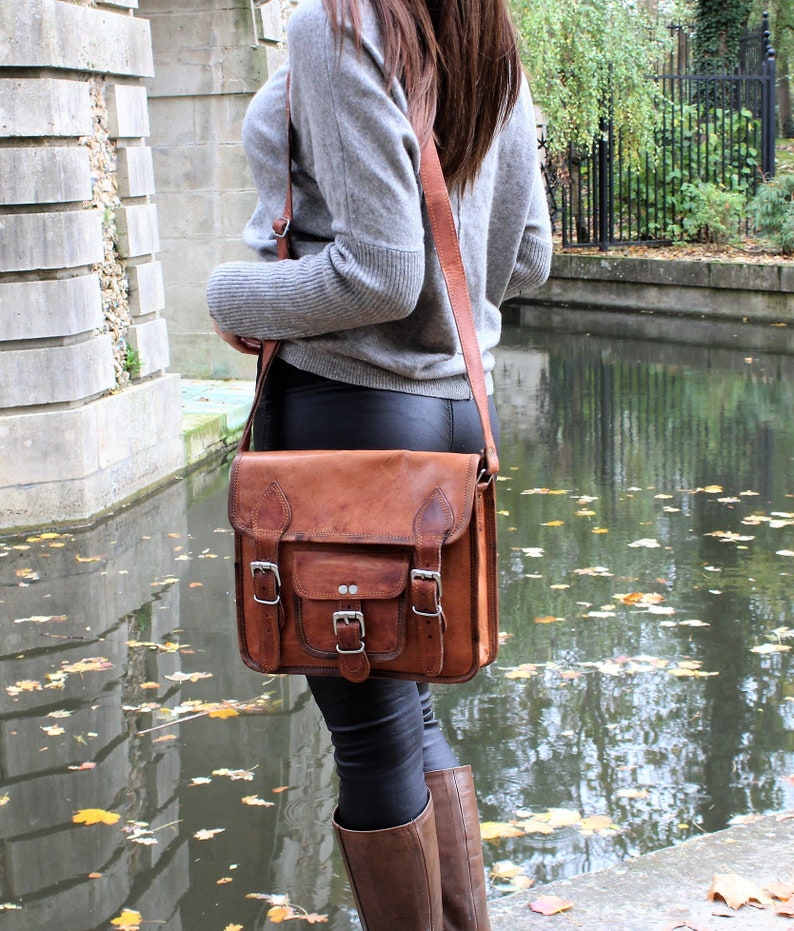 May include: A brown leather satchel bag with a flap closure and multiple buckles. The bag is worn over the shoulder of a person wearing a grey sweater and black trousers.