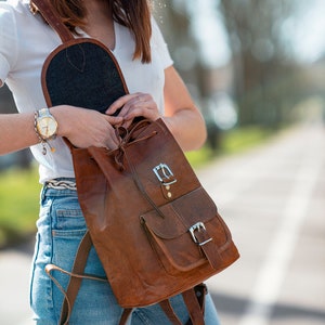 May include: A brown leather backpack with a flap and two front pockets, secured with silver buckles. The backpack has adjustable straps and a drawstring closure. The interior is lined with a dark fabric.