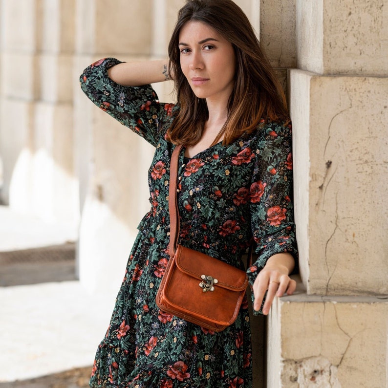 Op de afbeelding: Een bruine leren crossbodytas met een klepsluiting en een decoratieve sluiting. De tas heeft een lange, verstelbare riem. De vrouw draagt een jurk met bloemenprint met rode en groene bloemen.