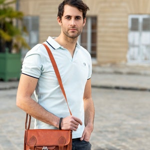 May include: A man wearing a light blue polo shirt with a brown leather satchel bag slung over his shoulder. The bag has two buckles and a flap with a pocket.