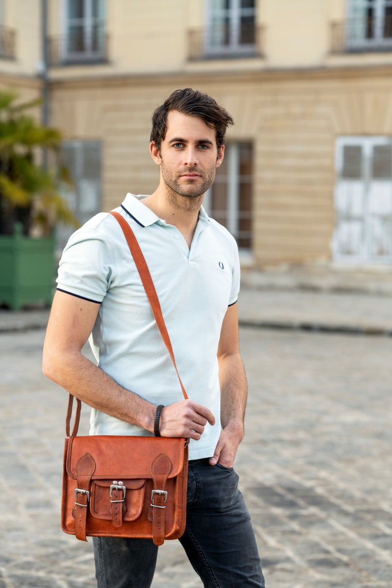 May include: A man wearing a light blue polo shirt with a brown leather satchel bag slung over his shoulder. The bag has two buckles and a flap with a pocket.