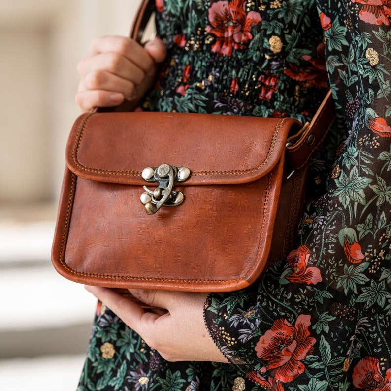 Op de afbeelding: Bruine leren crossbodytas met een rechthoekige vorm en een klepsluiting. De tas heeft een decoratieve metalen sluiting en een gestikte rand. De riem is verstelbaar. De tas wordt vastgehouden door een persoon die een bloemenjurk draagt.