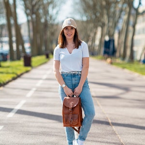 May include: A woman wearing a tan baseball cap with a white "LA" logo, a white v-neck t-shirt, blue jeans, and white sneakers. She is holding a brown leather backpack with buckles. The background features a tree-lined path.