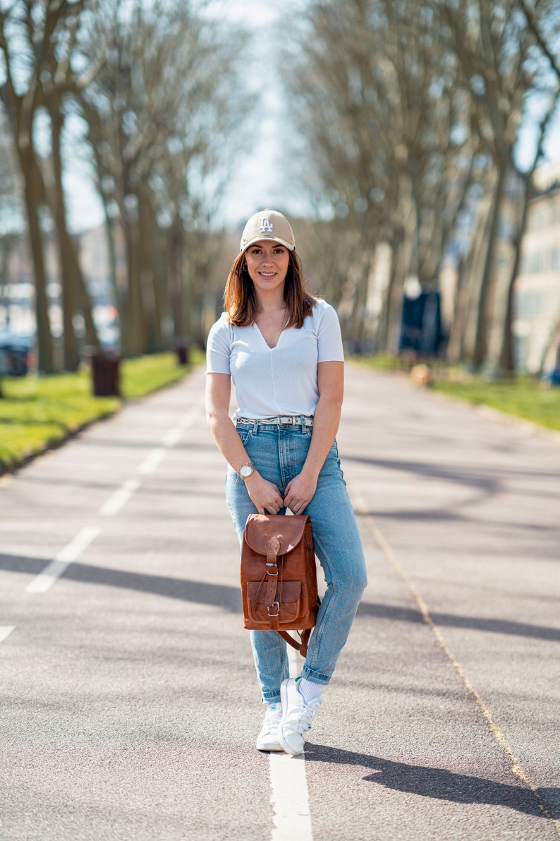 May include: A woman wearing a tan baseball cap with a white "LA" logo, a white v-neck t-shirt, blue jeans, and white sneakers. She is holding a brown leather backpack with buckles. The background features a tree-lined path.