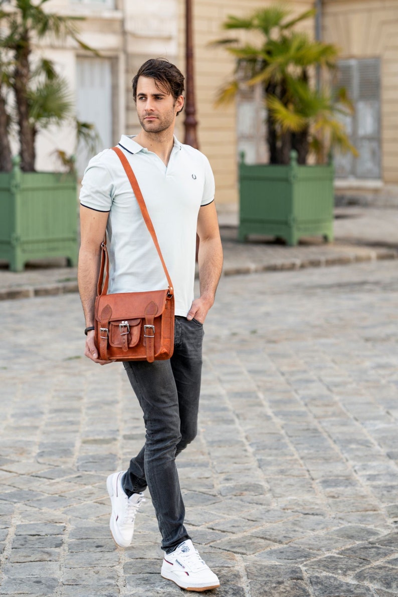 May include: A man wearing a light blue polo shirt with a brown leather satchel bag slung over his shoulder. He is wearing dark blue jeans and white trainers.