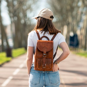 May include: A brown leather backpack with adjustable straps and a buckle closure. The backpack has a front pocket and is worn by a person wearing a white t-shirt, blue jeans, and a tan baseball cap.