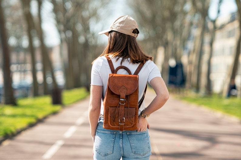 May include: A brown leather backpack with adjustable straps and a buckle closure. The backpack has a front pocket and is worn by a person wearing a white t-shirt, blue jeans, and a tan baseball cap.