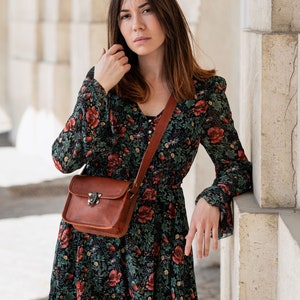 Op de afbeelding: Een vrouw draagt een zwarte jurk met bloemenprint met rode bloemen en een bruine leren crossbodytas. De tas heeft een metalen sluiting. De jurk heeft lange mouwen en een V-hals.