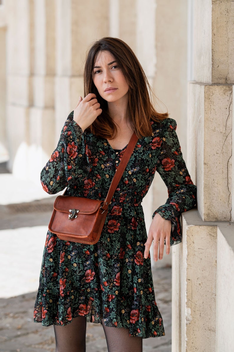 Op de afbeelding: Een vrouw draagt een zwarte jurk met bloemenprint met rode bloemen en een bruine leren crossbodytas. De tas heeft een metalen sluiting. De jurk heeft lange mouwen en een V-hals.