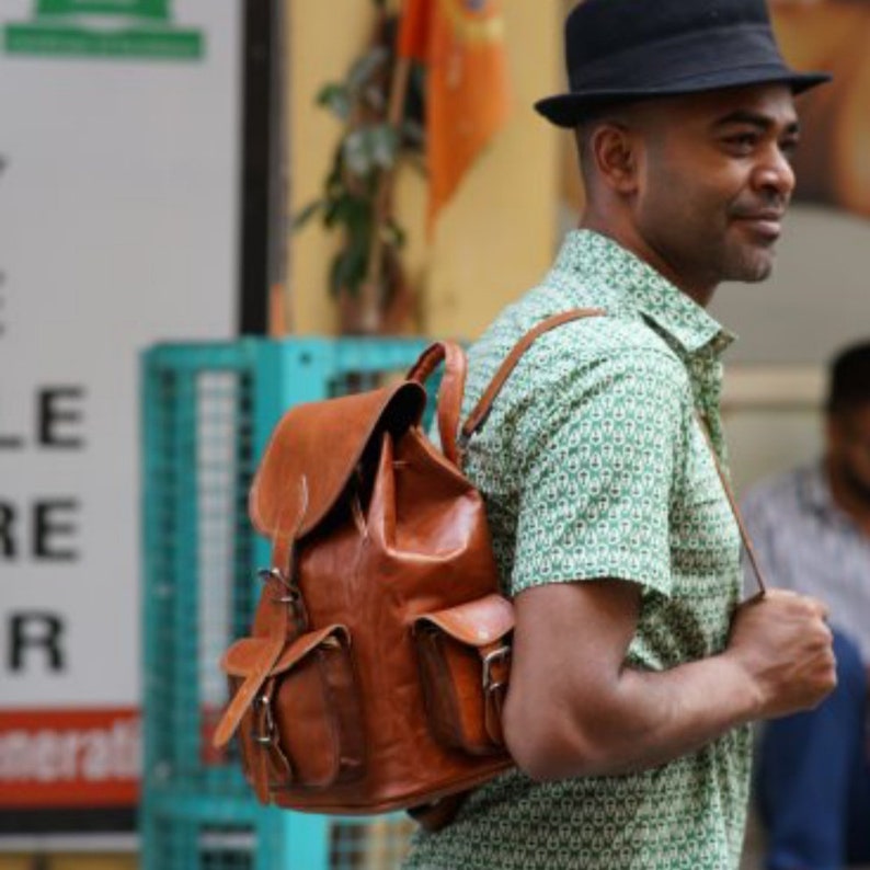 May include: A brown leather backpack with two front pockets and buckle closures. The backpack has adjustable straps and a flap top. The bag is worn by a person wearing a green patterned shirt and a black hat.