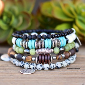 Könnte beinhalten: Ein Stapel Armbänder mit Perlen in verschiedenen Farben und Texturen. Die Armbänder umfassen schwarze, weiße, türkisfarbene, grüne, braune und gesprenkelte Perlen mit silberfarbenen Metallakzenten und einem Anhänger. Die Armbänder sind auf einer Holzoberfläche gestapelt.