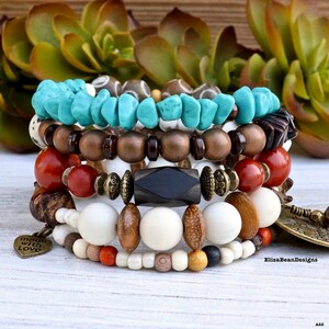 Könnte beinhalten: Gestapelte Armbänder mit türkisfarbenen, hölzernen und Steinperlen. Die Armbänder weisen eine Vielzahl von Farben und Texturen auf, mit einem herzförmigen Anhänger mit der Aufschrift "made with Love".