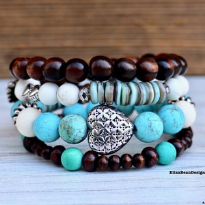 Puede incluir: Un conjunto de cinco pulseras apiladas, que incluyen cuentas de madera marrón, cuentas blancas, cuentas turquesa y un dije de corazón plateado. Las pulseras están apiladas sobre una superficie de madera blanca.