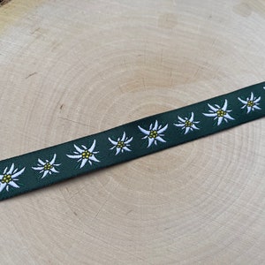 Puede incluir: Cinta verde oscuro con un patrón repetido de flores de edelweiss blancas y amarillas. La cinta se muestra en diagonal sobre una superficie de madera marrón claro, destacando los detalles y el contraste de color del diseño floral.