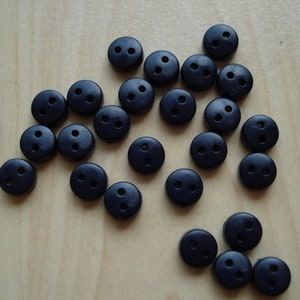 Może przedstawiać: Zbiór czarnych guzików z dwoma otworami. Guziki są rozrzucone na jasnym drewnianym podłożu.