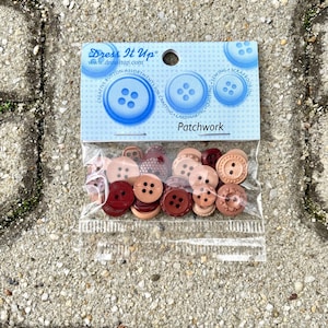 Puede incluir: Una bolsa de botones surtidos en tonos marrones y rojos. Los botones están en una bolsa de plástico transparente con una etiqueta que dice "Dress It Up" y "Patchwork".