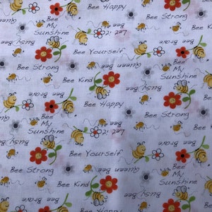Op de afbeelding: Witte stof met een herhalend patroon van bijen, bloemen en handschrift. De tekst bevat zinnen als "Bee Happy", "Bee Yourself", "Bee Strong" en "Bee Kind".