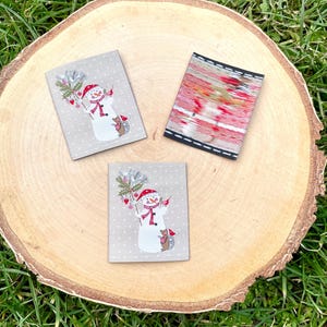 Peut inclure: Deux patchs en tissu beige avec un motif de bonhomme de neige et un patch en tissu à motifs rouge et blanc. Les patchs sont sur une surface en bois.
