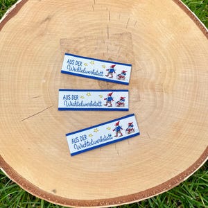 Op de afbeelding: Drie blauwe en witte stoffen labels met de tekst "AUS DER Wichtelwerkstatt" en een cartoon afbeelding van drie elven op een slee. De labels liggen op een houten oppervlak.