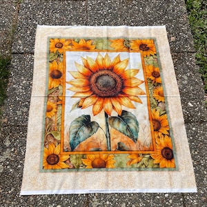 Può includere: Un pannello di tessuto rettangolare raffigurante un grande girasole centrale con petali gialli e arancioni e foglie verde scuro. L'immagine centrale è incorniciata da un bordo di girasoli più piccoli su uno sfondo beige chiaro con motivo.
