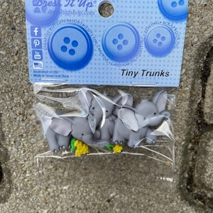 Könnte beinhalten: Eine Packung grauer, elefantenförmiger Knöpfe mit gelben Blumenakzenten. Die Verpackung ist mit "Tiny Trunks" beschriftet und enthält die Website-Adresse des Unternehmens sowie Social-Media-Symbole.