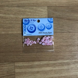 Może przedstawiać: Opakowanie różowych guzików w kształcie serca z napisem "MM Hearts Rose" od Dress It Up. Opakowanie zawiera liczne małe, jasno różowe guziki w kształcie serca. Opakowanie zawiera również niebieskie guziki i ikony mediów społecznościowych.