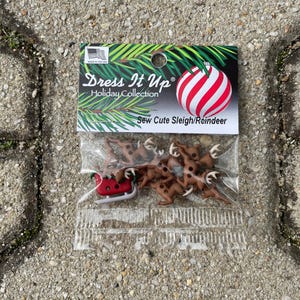 Könnte beinhalten: Eine Packung mit braunen Rentier- und Schlitten-Nähknöpfen. Die Packung ist mit "Dress It Up Holiday Collection Sew Cute Sleigh/Reindeer" beschriftet.