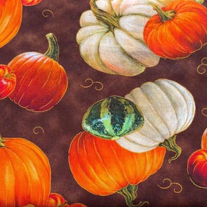 Op de afbeelding: Een stofprint met een herhalend patroon van oranje en witte pompoenen, kleine rode appels en groene kalebassen tegen een donkerbruine achtergrond. Het ontwerp bevat krullende ranken verspreid tussen de verschillende soorten oogstfruit.