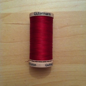 Könnte beinhalten: Eine Spule roten Gütermann-Baumwollgarns zum Quilten. Die Spule ist mit "Gütermann" und "Cotton Quilting" beschriftet.