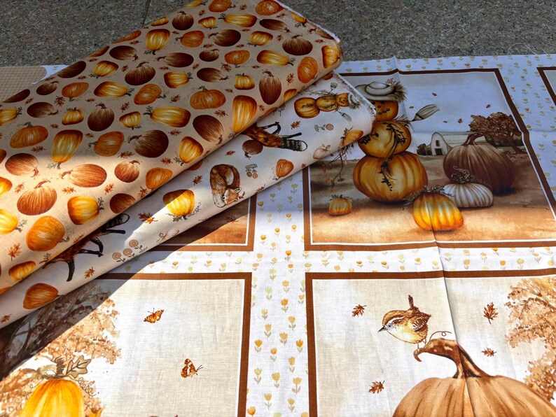 K&ouml;nnte beinhalten: Stoffrollen mit herbstlichen Mustern. Eine Rolle zeigt K&uuml;rbisse in Orange- und Braunt&ouml;nen auf beigem Hintergrund. Eine andere zeigt eine Herbstszene mit K&uuml;rbissen, einer Vogelscheuche und einem Bauernhaus. Der dritte Stoff hat ein K&uuml;rbis- und Vogeldesign.