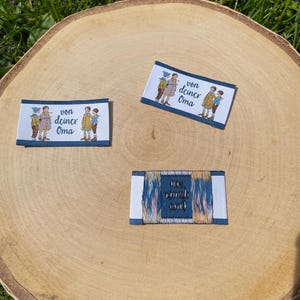 Könnte beinhalten: Drei blaue und weiße Stoff-Etiketten mit gesticktem Text und Bildern. Das erste Etikett lautet "von deiner Oma" und zeigt eine Comic-Abbildung von drei Kindern. Das zweite Etikett lautet "von deiner Oma" und zeigt eine Comic-Abbildung von drei Kindern. Das dritte Etikett lautet "100% recycled card" und zeigt ein farbenfrohes abstraktes Design.