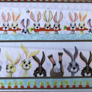 Op de afbeelding: Een witte stof met een herhalend patroon van konijnen. De konijnen staan in twee rijen, waarbij de bovenste rij konijnen laat zien die wortels vasthouden en de onderste rij konijnen laat zien met hun koppen die over een bruine lijn kijken. De konijnen zijn allemaal in verschillende kleuren, waaronder grijs, bruin en wit.