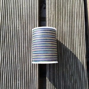 Può includere: Un'bobina di filo colorato con un'anima di plastica bianca. Il filo è a righe con sfumature di blu, verde, giallo e nero.