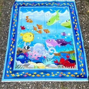 Könnte beinhalten: Ein farbenfroher Stoff mit einer Unterwasserszene mit Cartoon-Fischen, einer Krabbe und einem Oktopus. Das Design umfasst einen blauen Rand mit kleinen gelben Fischen. Die Hauptszene hat einen blauen Hintergrund mit verschiedenen Fischen in Orange-, Grün-, Lila- und Gelbtönen.