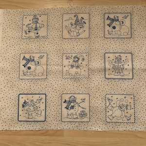 Könnte beinhalten: Ein beigefarbener Stoff mit einem sich wiederholenden Muster aus neun blauen Quadraten. Jedes Quadrat zeigt ein anderes Schneemann-Design mit verschiedenen Winteraktivitäten, darunter Skifahren, Schneemann bauen und einen Vogel halten.