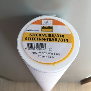 Peut inclure: Un rouleau de tissu non tissé STICKVLIES/314 STITCH-N-TEAR/314 blanc. L'étiquette est orange et jaune avec la marque "vlieseline" et le logo Freudenberg. Le rouleau mesure 90 cm x 15 m, composé de 70% CV, 30% pâte de bois.