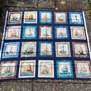 Op de afbeelding: Een marineblauw stoffen paneel met een herhalend patroon van ingelijste nautische scènes. Elke vierkant toont een andere illustratie van een zeilboot of vuurtoren, tegen een blauwgroene en bruine achtergrond. De stof is waarschijnlijk katoen of een vergelijkbaar materiaal.