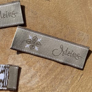Könnte beinhalten: Rechteckige Webetiketten in einem gedeckten Beigeton. Jedes Etikett zeigt links ein weiß gesticktes vierblättriges Blumenmotiv und rechts den in passendem beigen Garn gestickten kursiven Schriftzug "Meins".