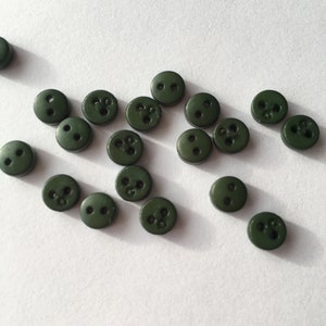 Op de afbeelding: Een verzameling donkergroene plastic knopen met twee of drie gaten. De knopen zijn verspreid over een wit oppervlak.
