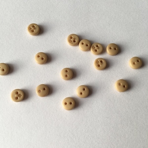 Peut inclure: Un ensemble de 14 boutons en plastique beige à deux trous chacun. Les boutons sont éparpillés sur une surface blanche.