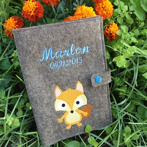 Puede incluir: Una carpeta de fieltro gris con cierre de bot&oacute;n azul. La carpeta tiene un dise&ntilde;o de zorro bordado y el texto "Marlon 04.11.2015" en hilo azul.