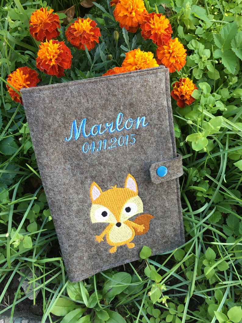 Puede incluir: Una carpeta de fieltro gris con cierre de bot&oacute;n azul. La carpeta tiene un dise&ntilde;o de zorro bordado y el texto "Marlon 04.11.2015" en hilo azul.