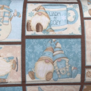 Könnte beinhalten: Ein blauer und brauner Stoff mit einem sich wiederholenden Muster aus Gnomen, Schneemännern und Schneeflocken. Der Stoff zeigt eine Tasse mit dem Text "Warm 'n Cozy".