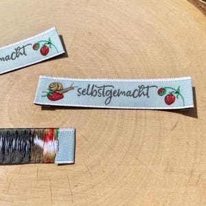 Puede incluir: Dos etiquetas de tela azul claro con costuras blancas. La etiqueta superior tiene la palabra "macht" bordada en negro. La etiqueta inferior tiene un caracol con una concha verde y un cuerpo rojo arrastrándose por una vid verde con fresas rojas. La etiqueta también tiene el texto "selbstgemacht" bordado en negro.