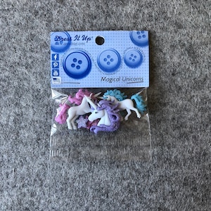 Op de afbeelding: Een pakketje "Magical Unicorns" versieringen. Het pakketje bevat kleine, witte eenhoornfiguren met kleurrijke manen in roze, paars en blauw. Het pakketje bevat ook een blauw en wit label met de woorden "Dress It Up".