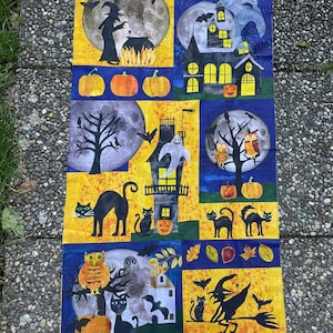 Puede incluir: Un panel de tela amarillo y azul con temática de Halloween. El panel presenta imágenes de gatos, búhos, brujas, fantasmas, calabazas y una casa encantada. El texto "Round Robin by MJ  Gunn" está impreso en la esquina inferior izquierda.