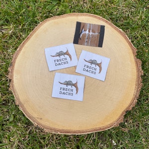 May include: Three white fabric labels with a brown badger illustration and the text "FRECH DACHS" on a wooden surface. A smaller brown and white fabric label is also visible.
