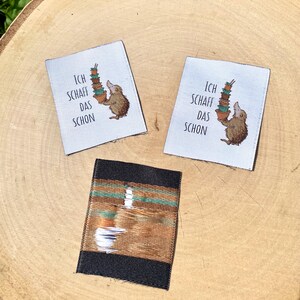 May include: Three fabric labels on a wooden surface. Two labels are white with a cartoon hedgehog carrying a stack of pots and the German text "ICH SCHAFF DAS SCHON". The third label is black with a brown and teal design.
